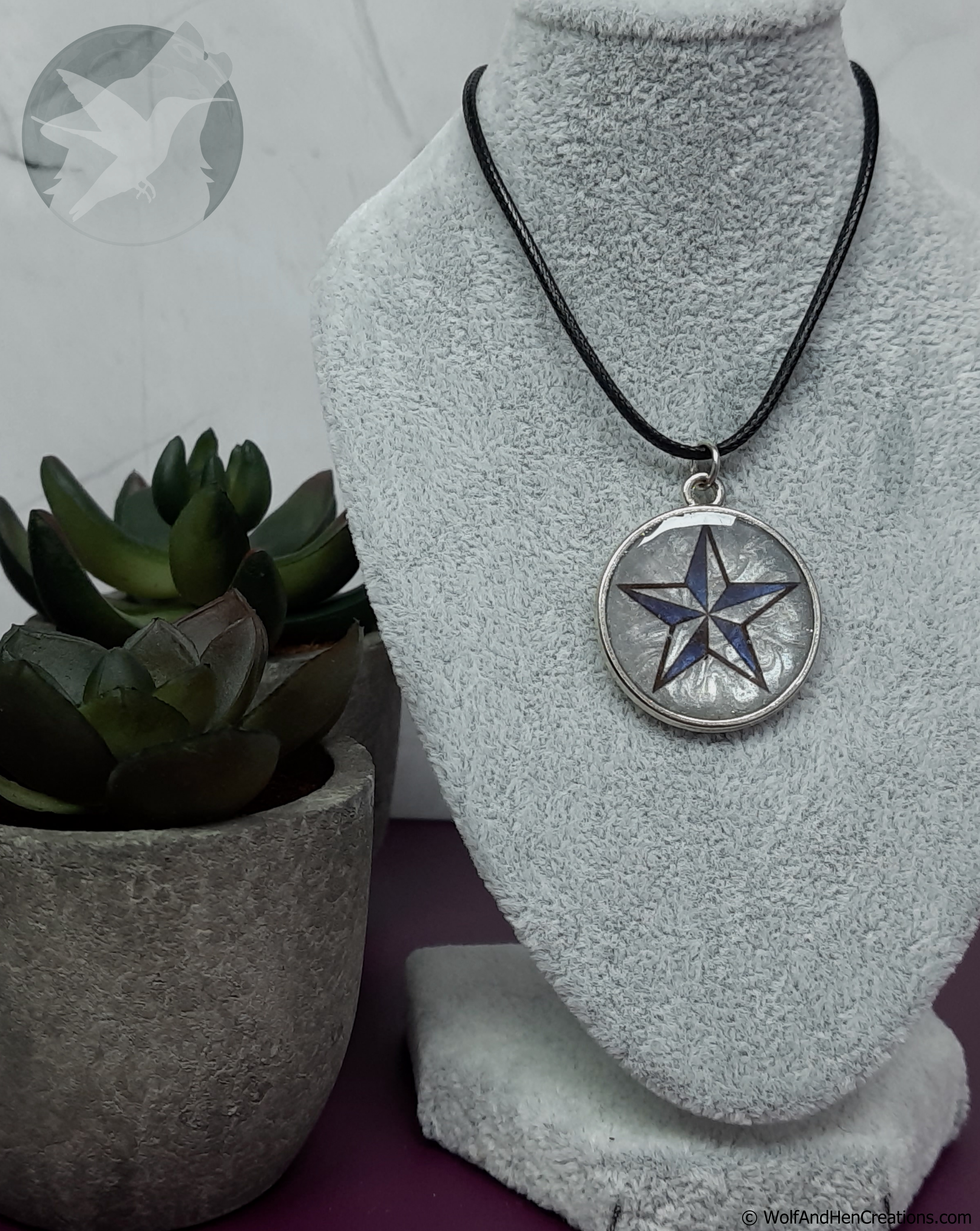 A silver circular pendant necklace featuring a blue and black five-pointed star design, displayed on a grey fabric jewellery bust with a small potted succulent in the background.