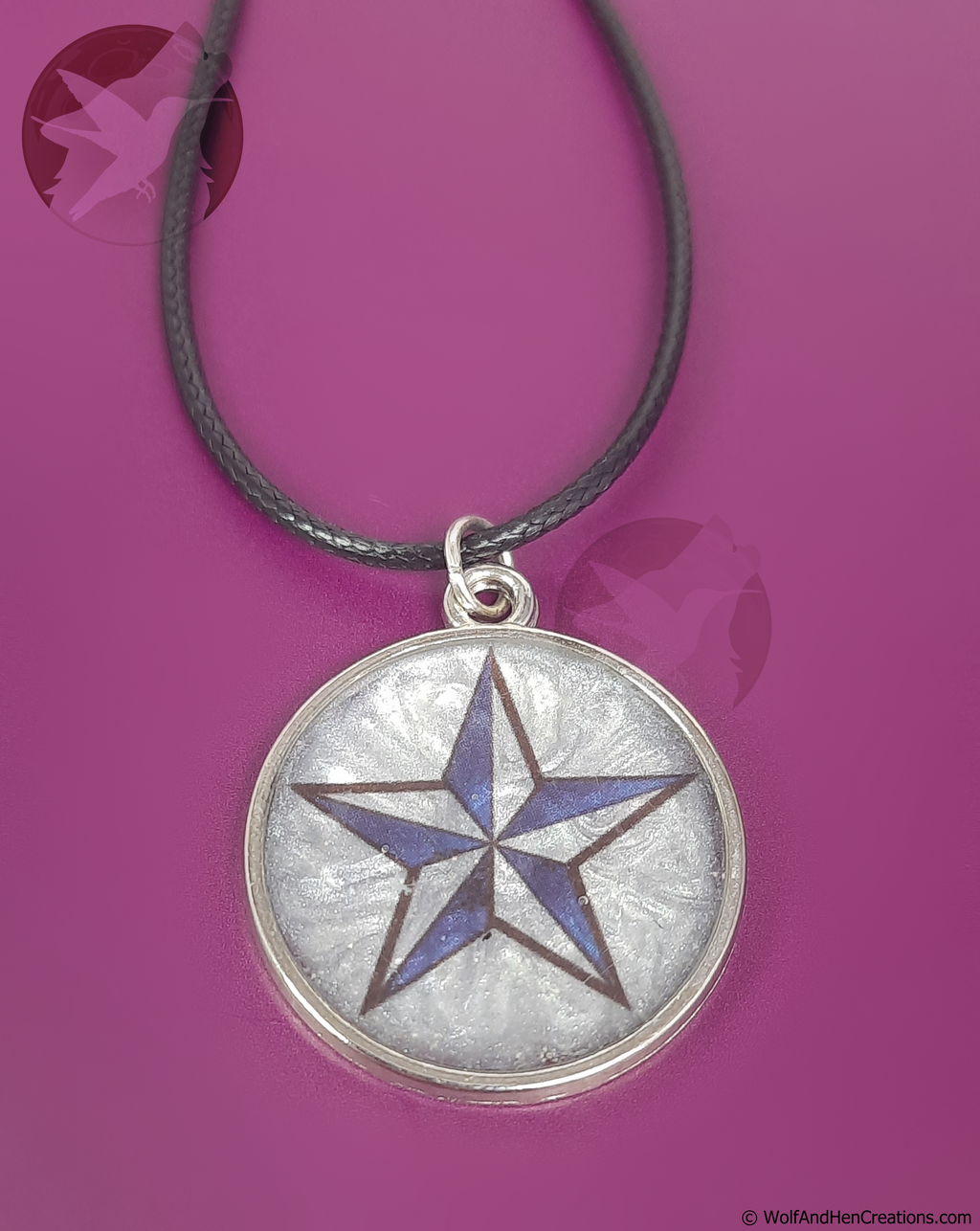 A circular pendant necklace featuring a silver-toned bezel with a silver textured inlay and a central blue and black star design, suspended from a black cord against a solid magenta background.