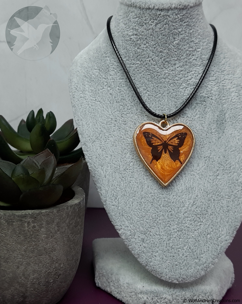 A heart-shaped pendant necklace featuring a butterfly design on orange swirled resin, displayed on a grey fabric bust. The pendant is suspended from a black cord and is edged in gold toned metal.