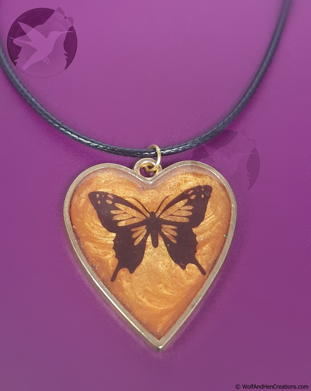 A heart-shaped, gold framed, pendant necklace featuring a black butterfly design on a textured orange resin background, suspended from a black cord against a solid purple background.