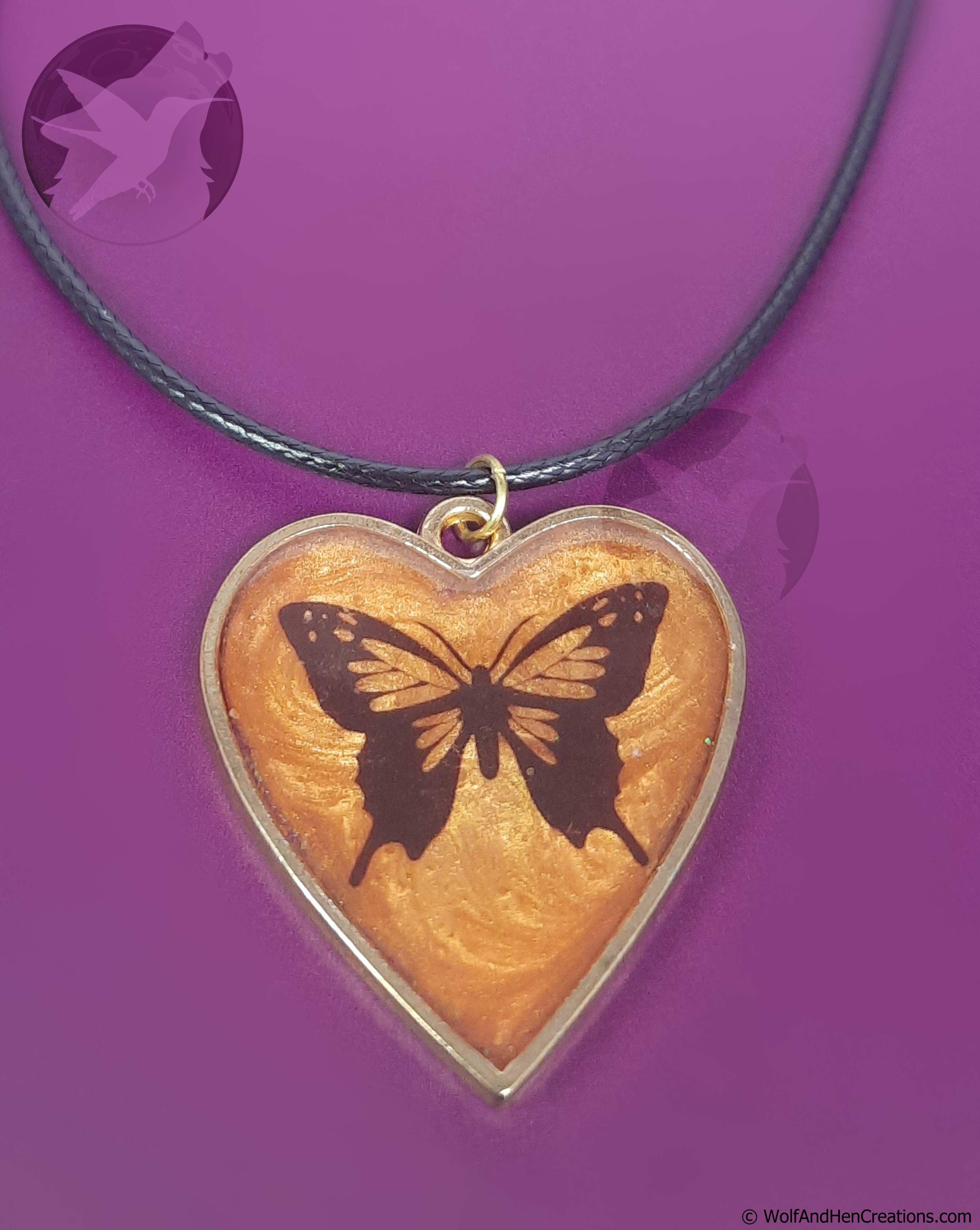 A heart-shaped, gold framed, pendant necklace featuring a black butterfly design on a textured orange resin background, suspended from a black cord against a solid purple background.