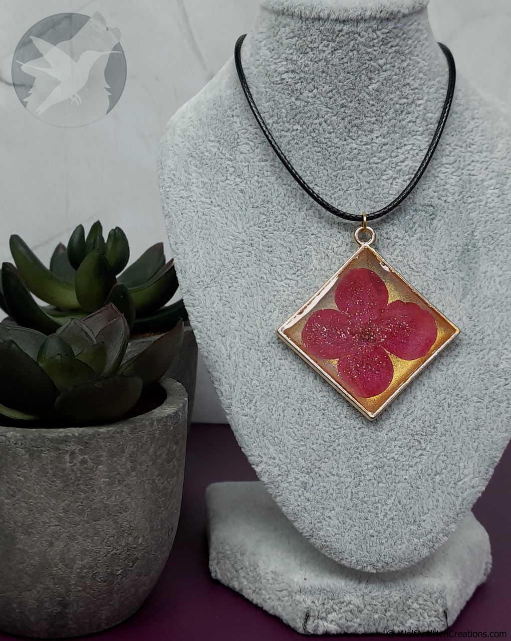 A square pendant necklace displayed on a grey fabric jewellery bust, featuring a pressed pink flower on a gold background with a sprinkle of glitter, on a black cord. A potted succulent is visible on the left.