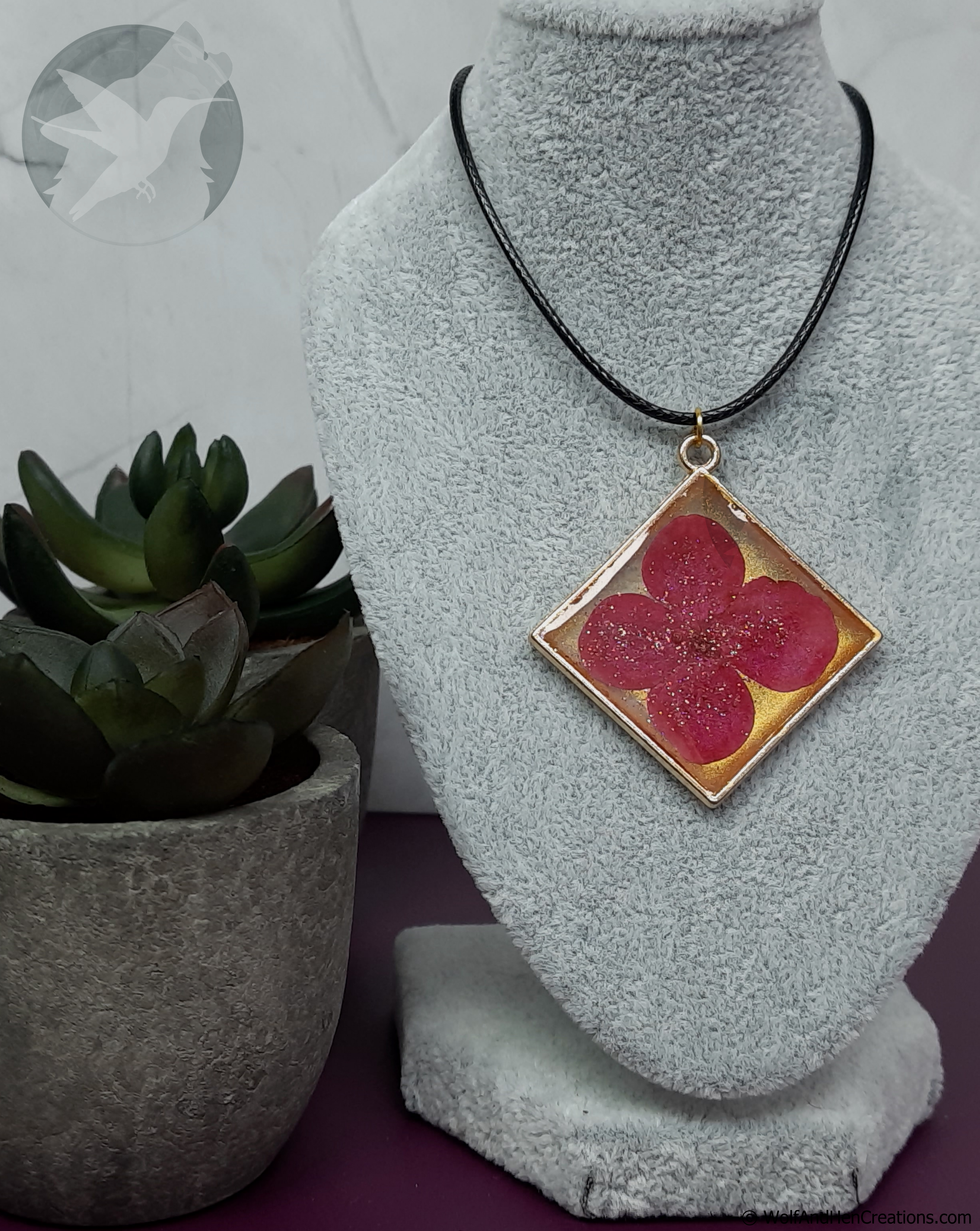 A square pendant necklace displayed on a grey fabric jewellery bust, featuring a pressed pink flower on a gold background with a sprinkle of glitter, on a black cord. A potted succulent is visible on the left.