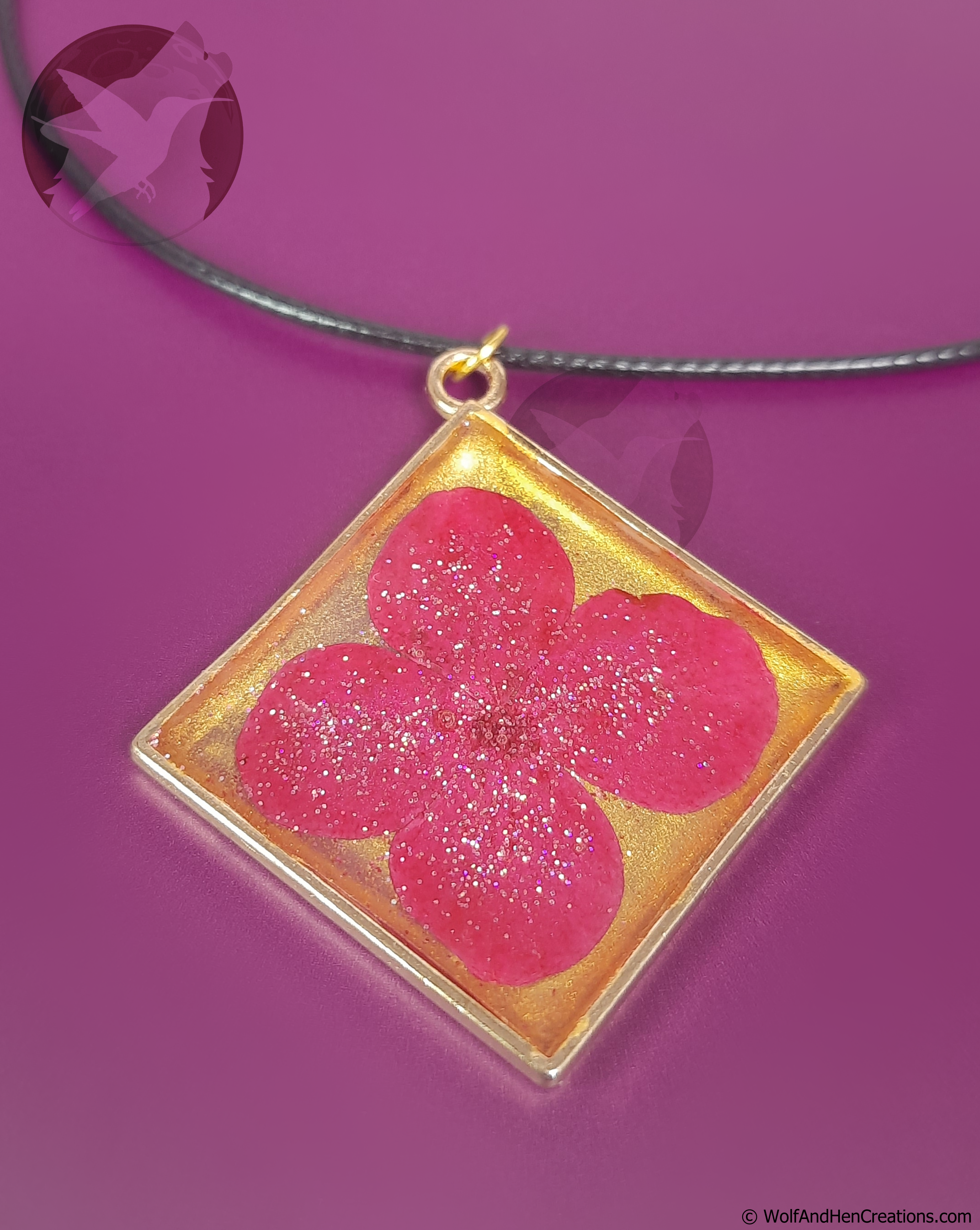 A square pendant necklace featuring a  pink pressed flower on a gold background set in a matching gold toned frame, suspended from a black cord against a solid purple background.