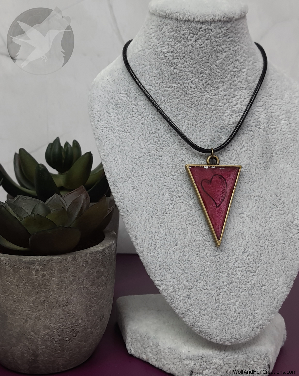 Necklace with a red triangle pendant featuring a heart design on a gray stand with a plant in the background.