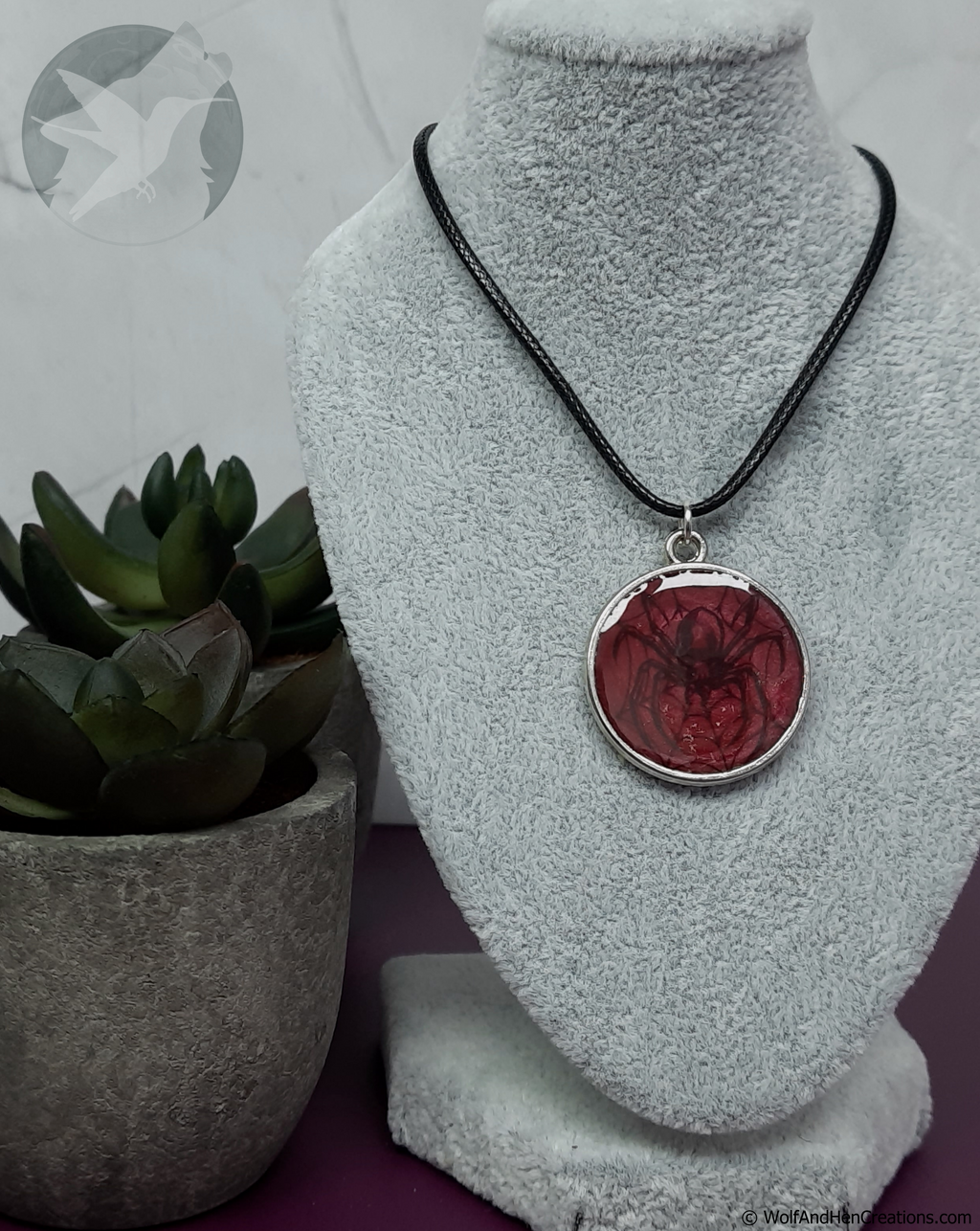 A round pendant necklace with a red spider design set in silver, hanging on a black cord from a gray velvet display bust, with a small potted succulent on the left.