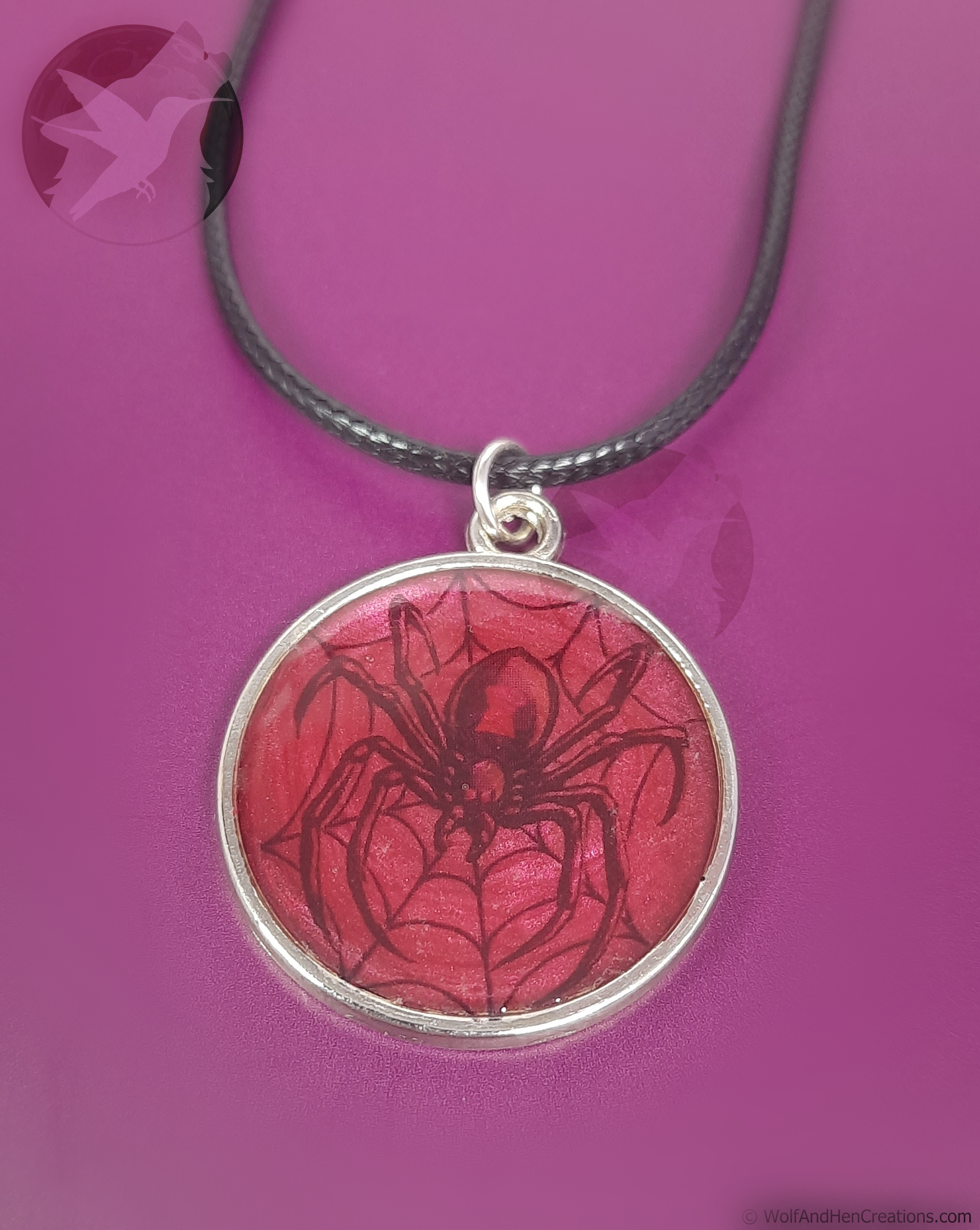 A round pendant necklace featuring a red spider web design with a spider in the center, set in a silver bezel on a dark gray cord, shown against a solid magenta background.