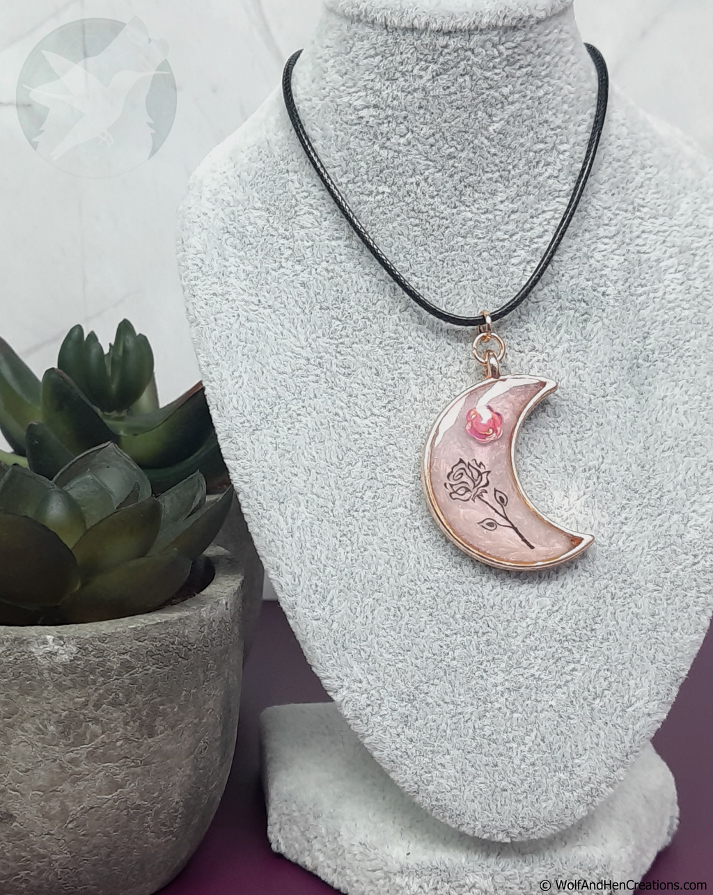 A crescent moon pendant necklace with pink and black rose designs and a rose gold border, displayed on a grey textured mannequin bust next to a small potted succulent.