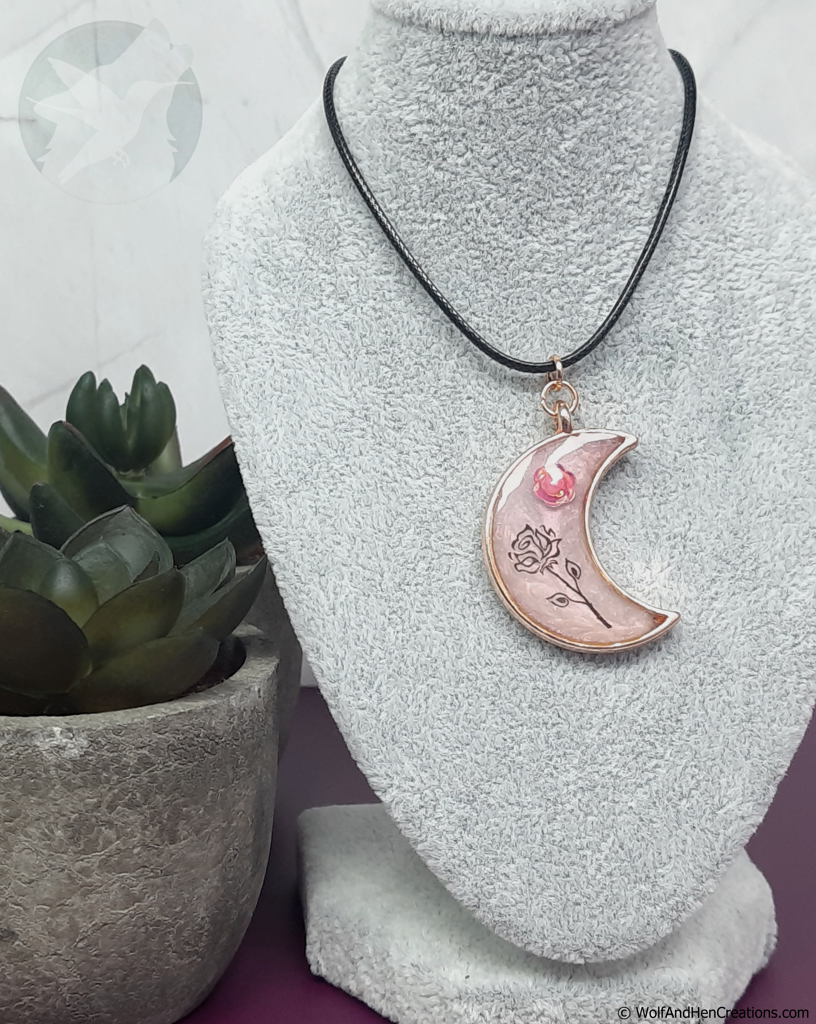 A crescent moon pendant necklace with pink and black rose designs and a rose gold border, displayed on a grey textured mannequin bust next to a small potted succulent.