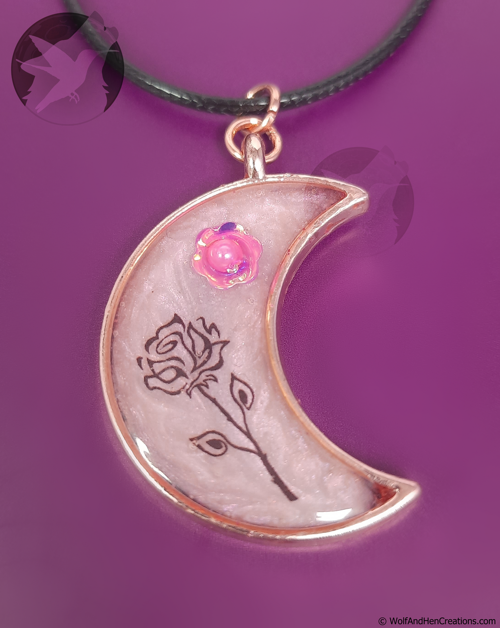A crescent moon pendant necklace with a rose pink background featuring a pink pearl flower charm and a black rose design, suspended on a black cord against a purple background.