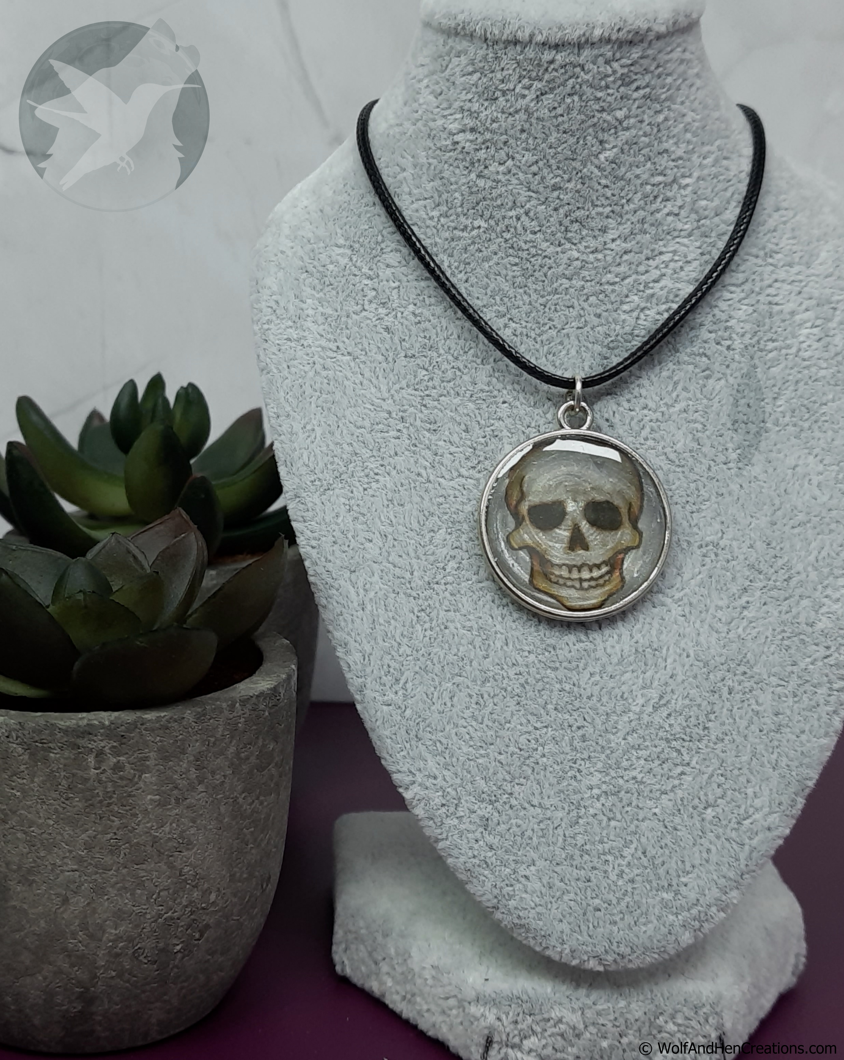 A silver pendant necklace featuring a detailed skull design, displayed on a gray velvet jewelry bust. The necklace is paired with a black cord, and the bust is styled next to a small potted succulent plant.