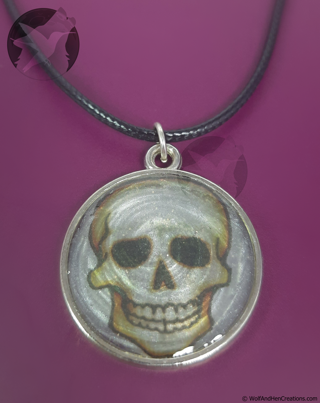A circular silver-toned pendant necklace featuring a painted skull design, suspended from a black cord against a solid magenta background.