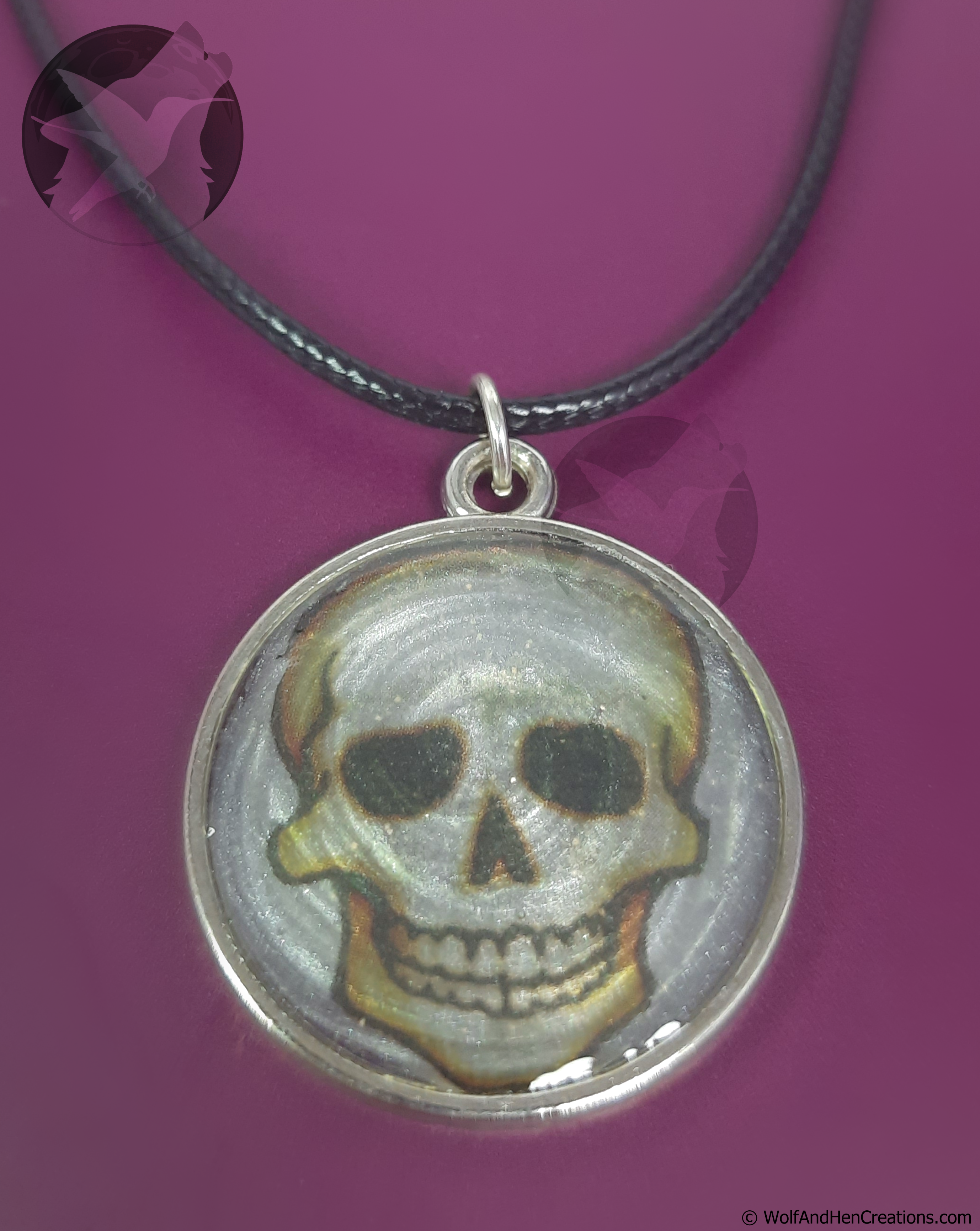 A circular silver-toned pendant necklace featuring a painted skull design, suspended from a black cord against a solid magenta background.