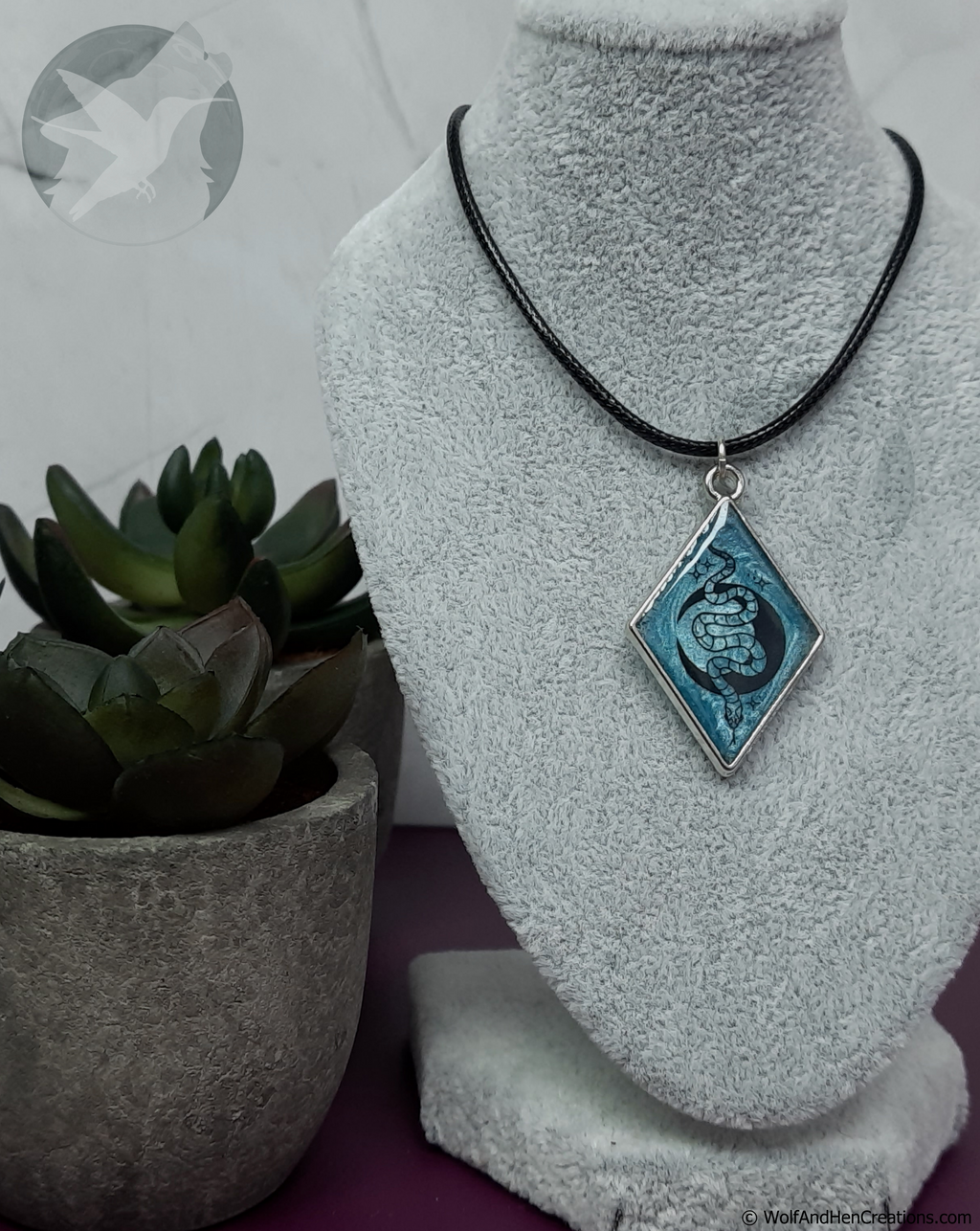 A diamond-shaped pendant necklace with a blue and black snake and moon design with a silver border, displayed on a grey velvet jewellery bust next to a small potted succulent.