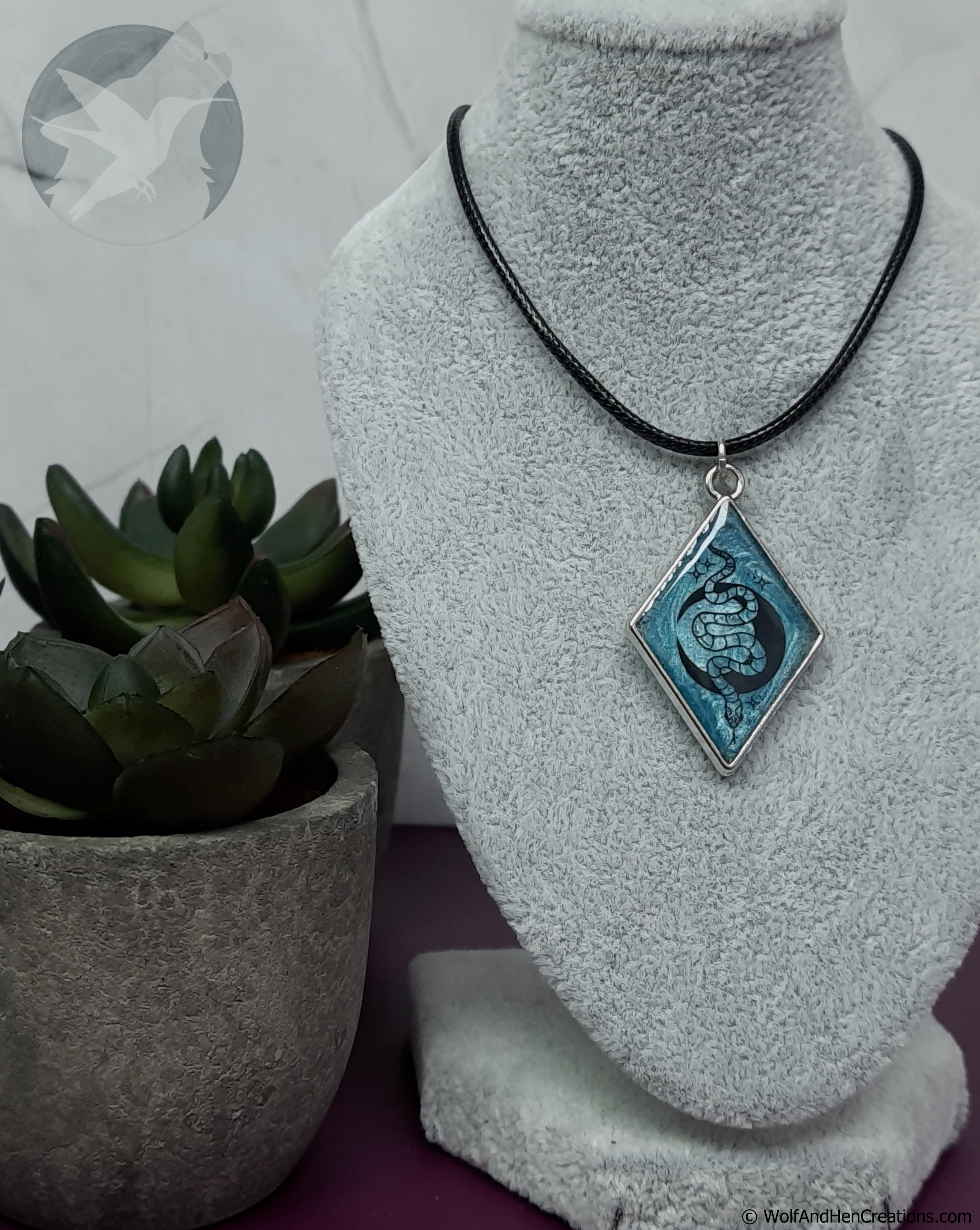 A diamond-shaped pendant necklace with a blue and black snake and moon design with a silver border, displayed on a grey velvet jewellery bust next to a small potted succulent.
