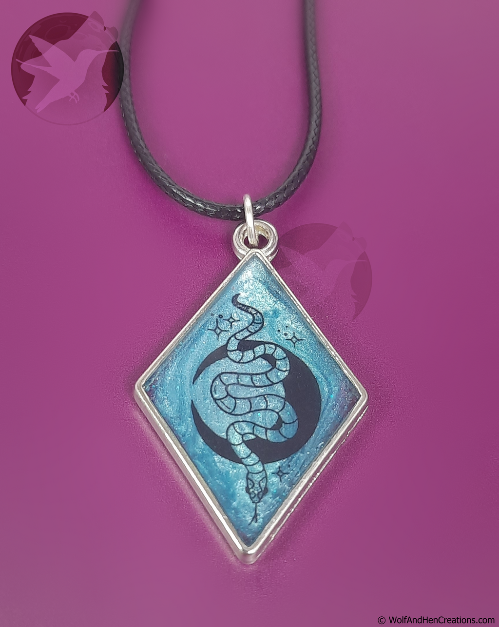 A diamond-shaped pendant necklace featuring a blue and black snake and moon design on a silver frame, suspended from a black cord against a vibrant magenta background.