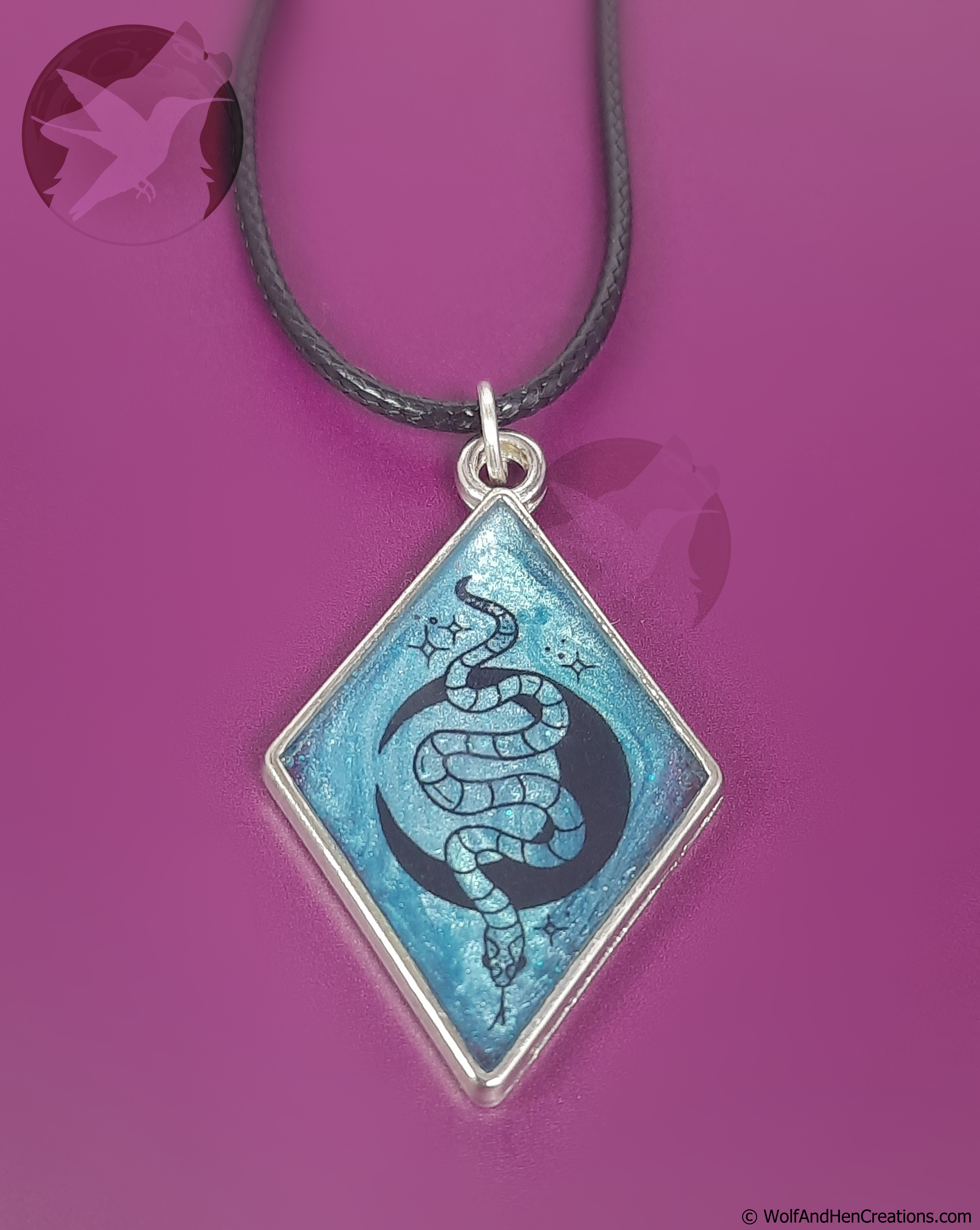 A diamond-shaped pendant necklace featuring a blue and black snake and moon design on a silver frame, suspended from a black cord against a vibrant magenta background.