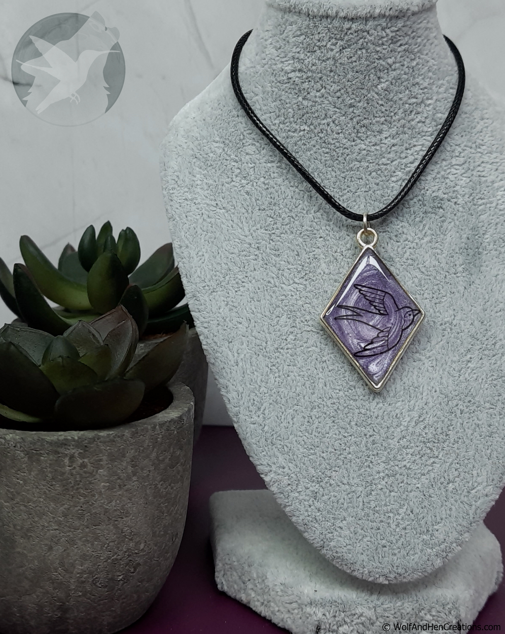 A diamond-shaped pendant necklace with a purple and black bird design, set in a pale gold tone frame and suspended from a black cord, displayed on a grey fabric jewellery bust next to a small potted succulent.