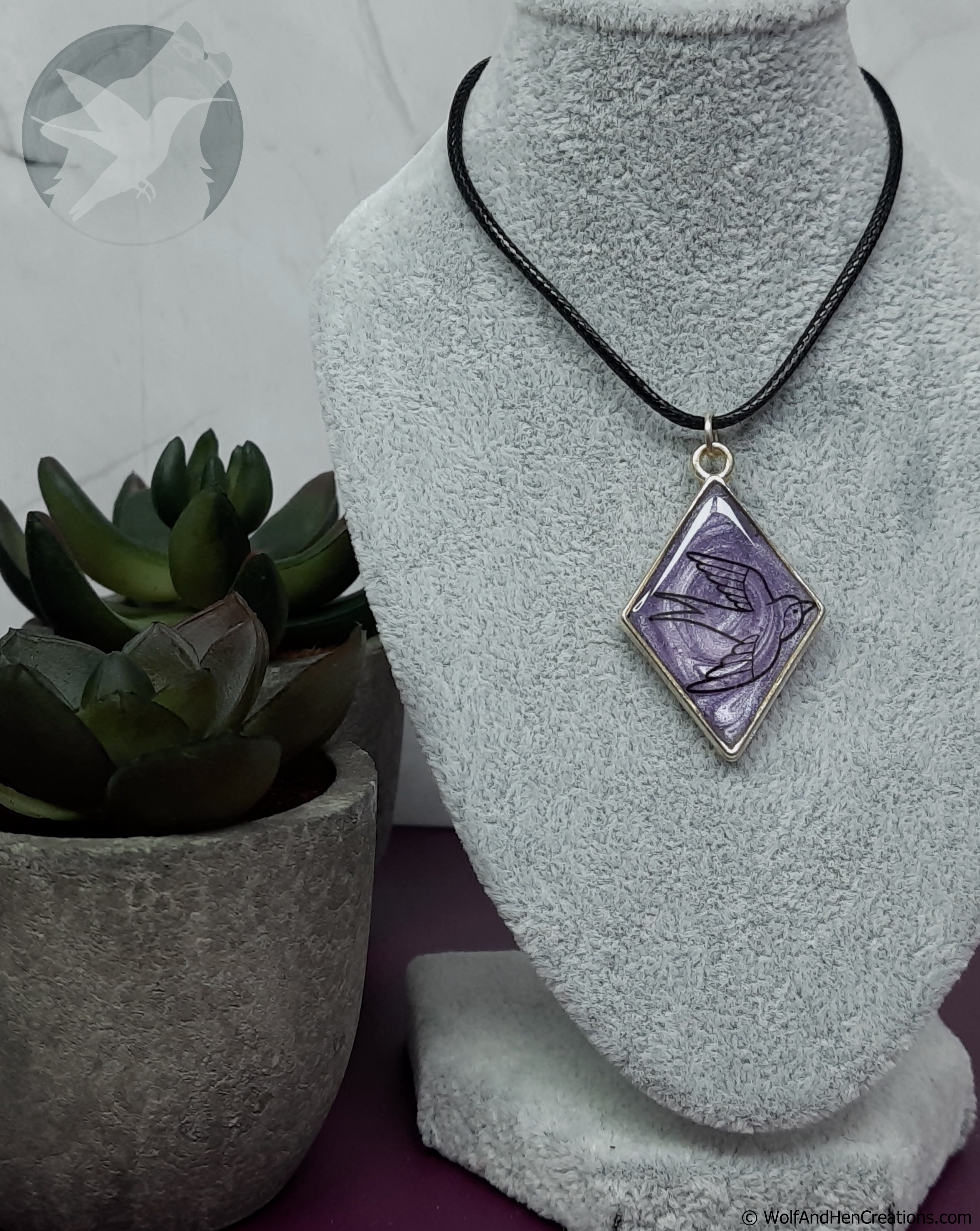 A diamond-shaped pendant necklace with a purple and black bird design, set in a pale gold tone frame and suspended from a black cord, displayed on a grey fabric jewellery bust next to a small potted succulent.