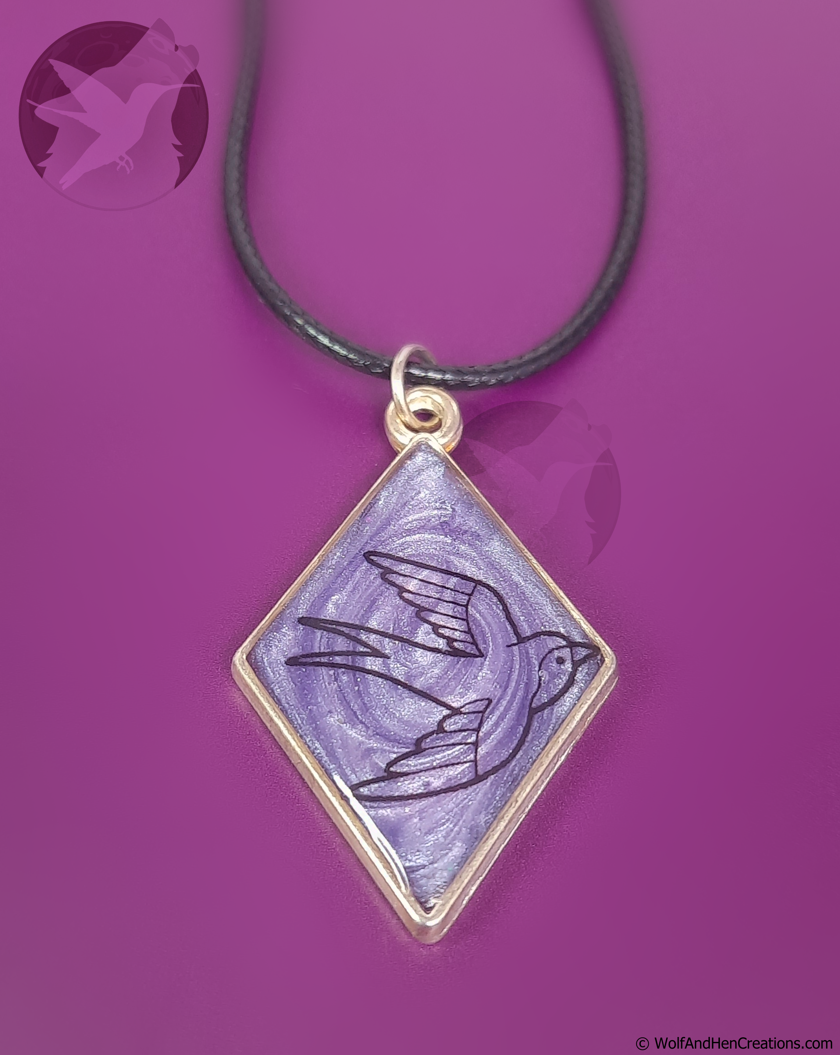 A diamond-shaped pendant necklace featuring a lilac background with a black bird illustration, set in a gold-tone frame and suspended from a black cord, shown against a solid purple background.