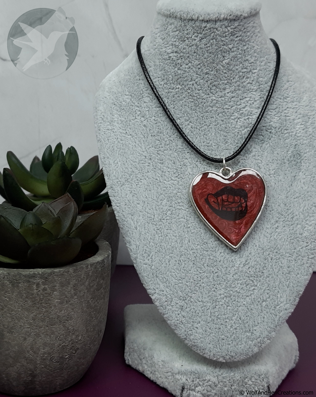 A heart-shaped pendant necklace with a red and black design, displayed on a grey fabric jewellery bust. The pendant features a silver-coloured border and a black graphic resembling a mouth with vampire teeth.