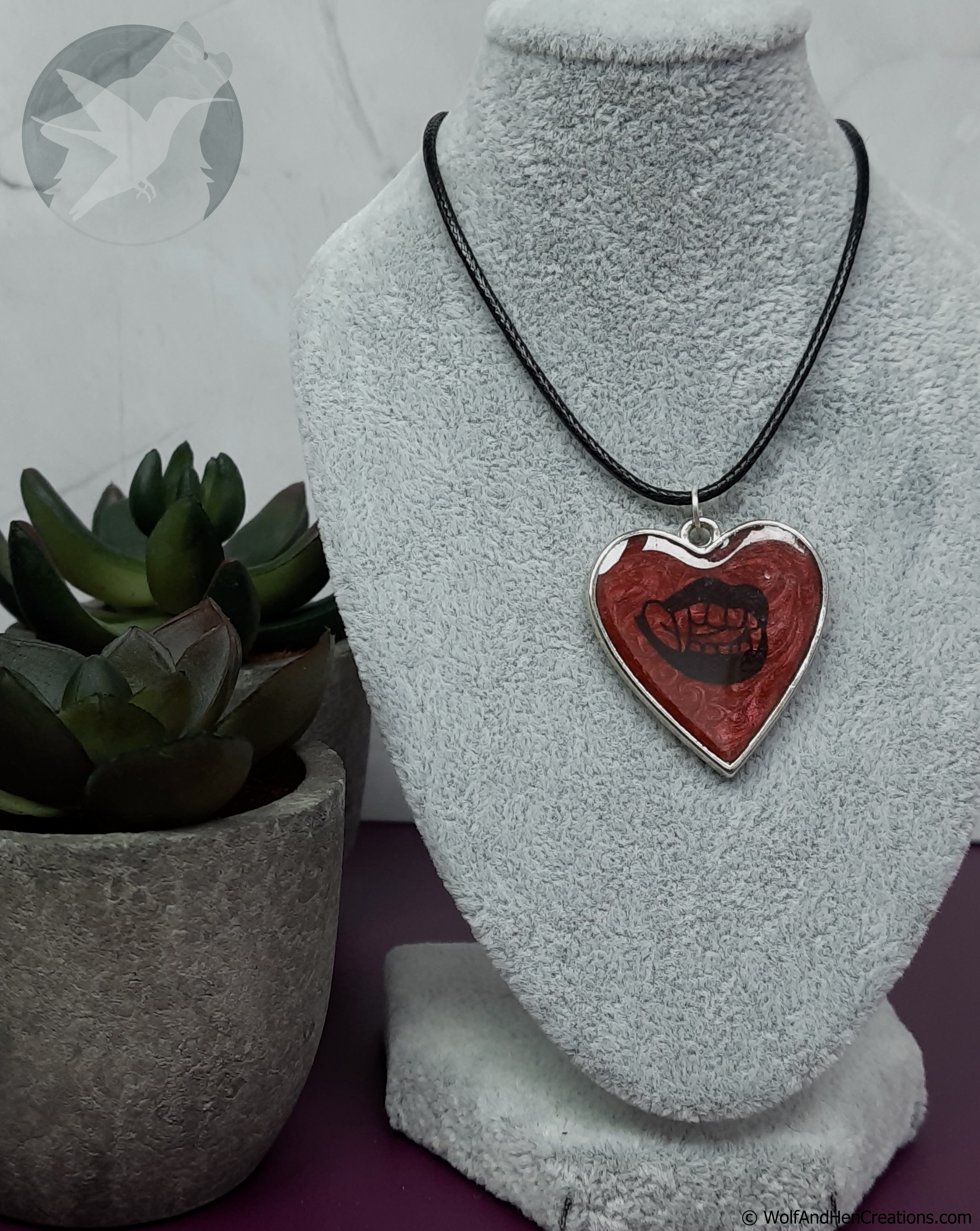 A heart-shaped pendant necklace with a red and black design, displayed on a grey fabric jewellery bust. The pendant features a silver-coloured border and a black graphic resembling a mouth with vampire teeth.