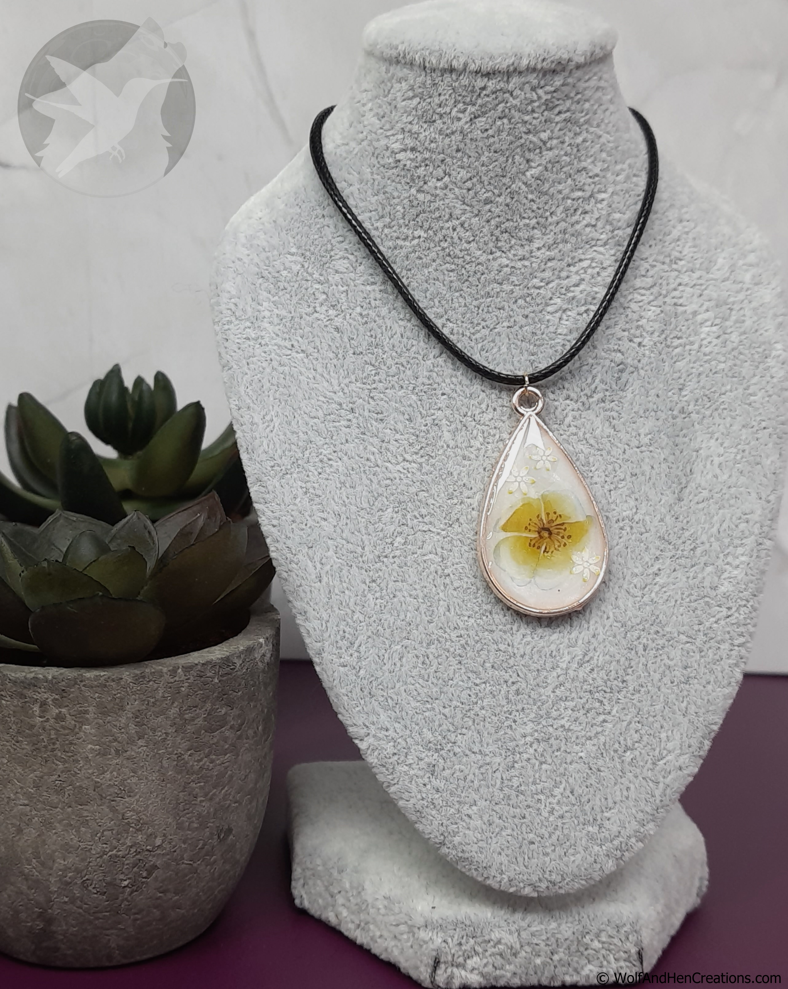 Teardrop-shaped pendant with floral design on a gray stand, next to a potted plant.