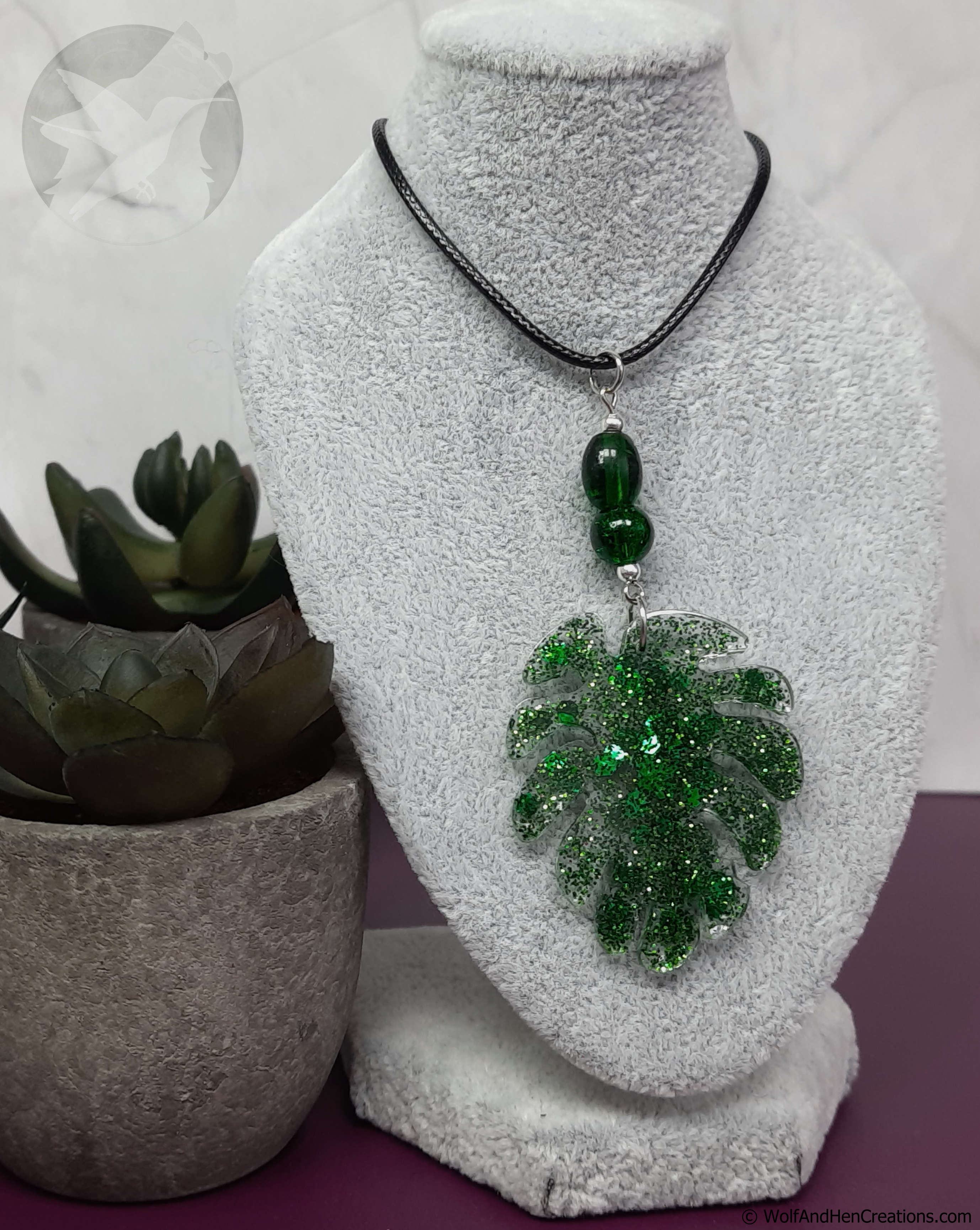 Green leaf-shaped pendant on a necklace displayed on a white bust with a plant in the background.