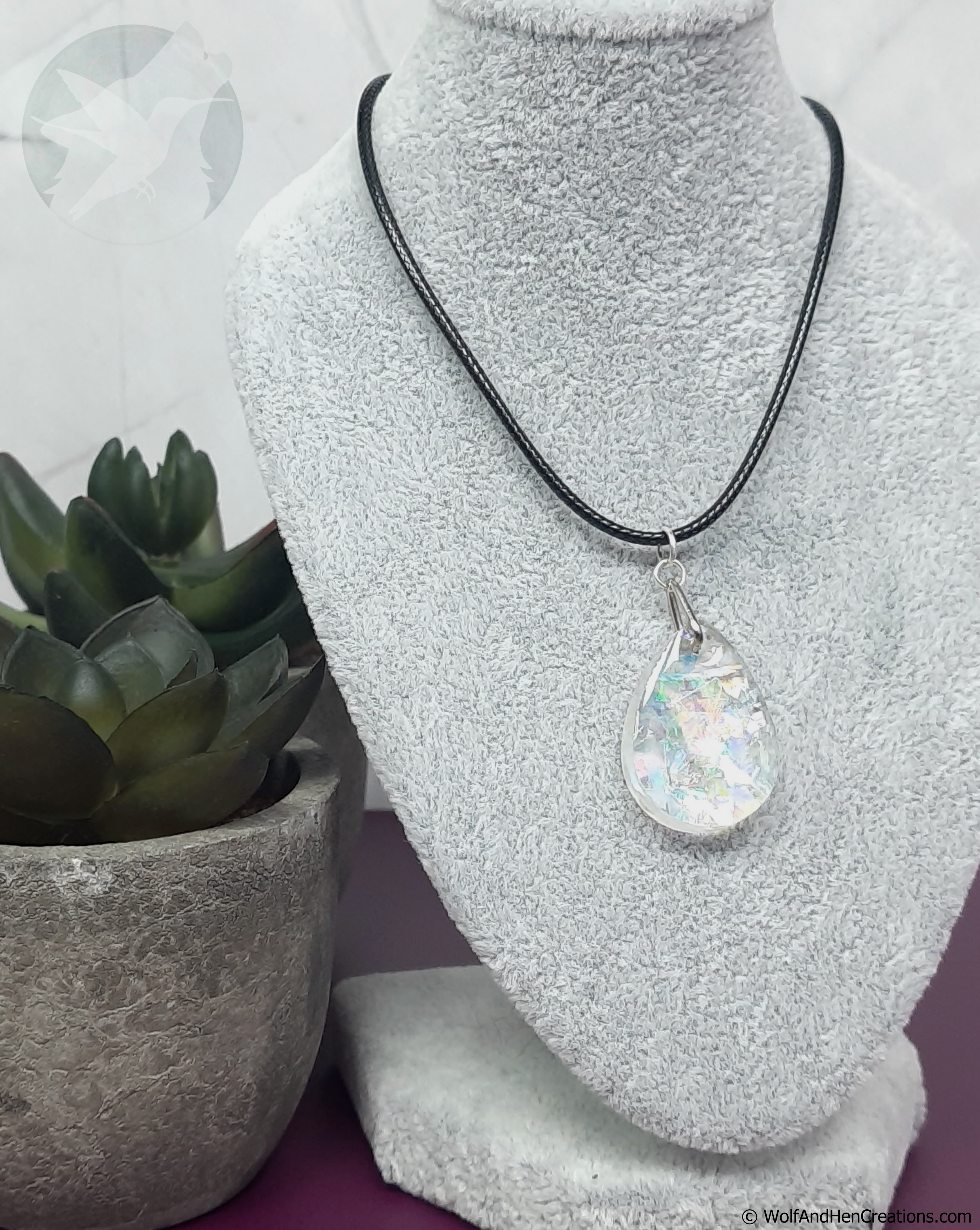 A teardrop shaped iridescent imitation opal pendant necklace on a black cord, displayed on a grey textured jewellery bust next to a small potted succulent.