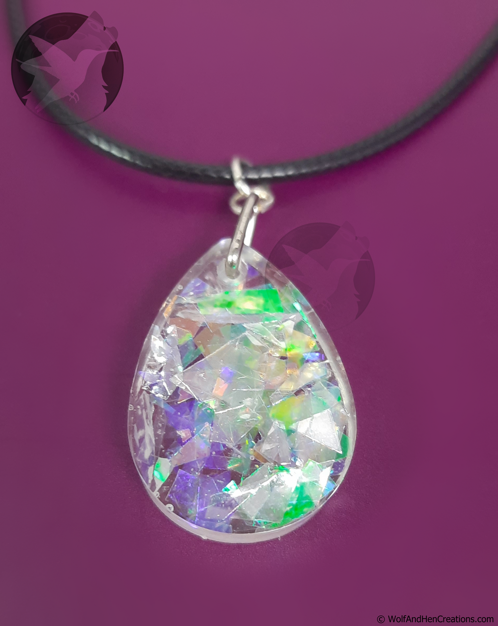 A teardrop shaped clear resin pendant filled with iridescent imitation opal facets that shift between green, purple, and gold, suspended from a black cord necklace against a solid purple background.