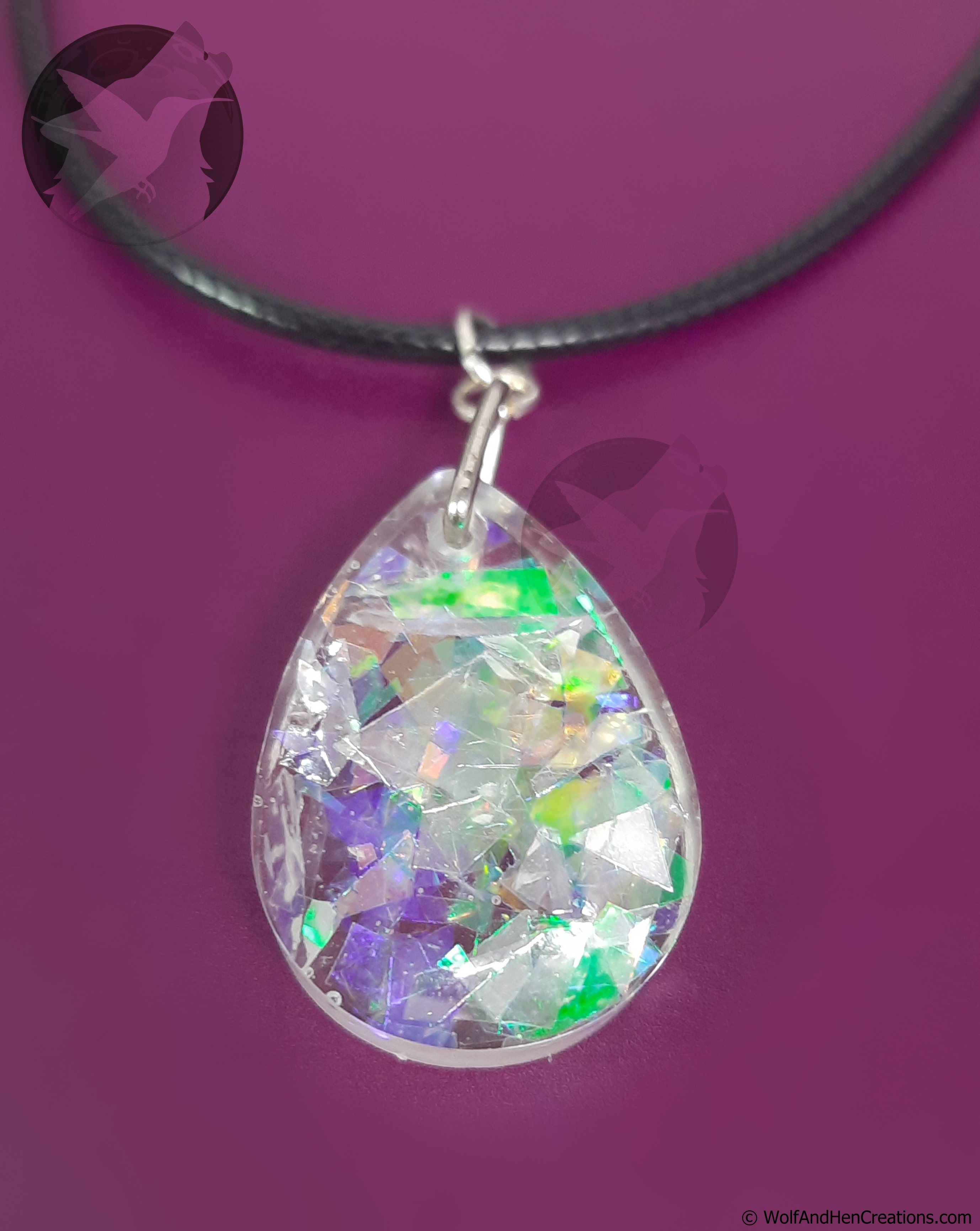 A teardrop shaped clear resin pendant filled with iridescent imitation opal facets that shift between green, purple, and gold, suspended from a black cord necklace against a solid purple background.