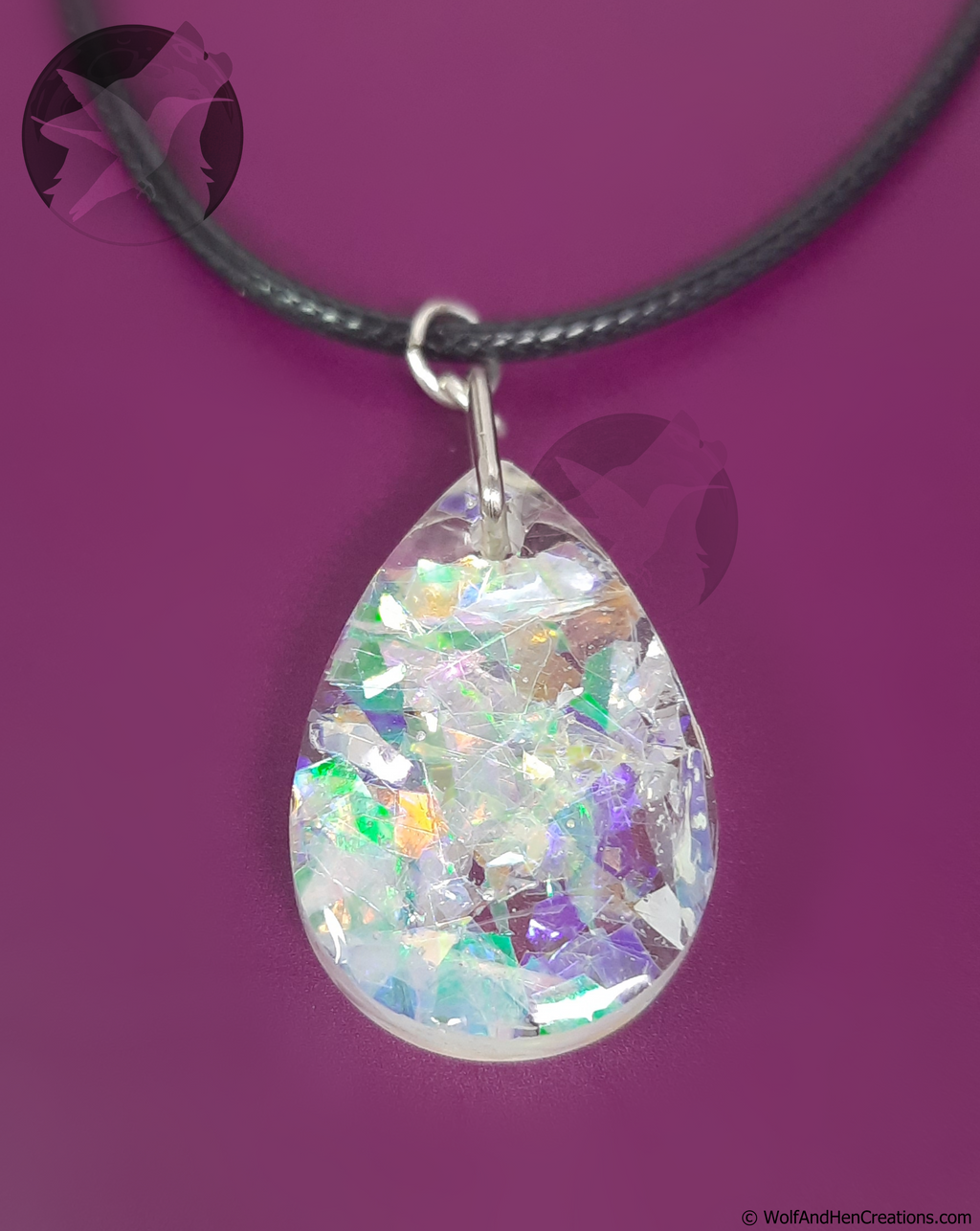 A teardrop shaped clear resin pendant filled with iridescent imitation opal facets, suspended from a black cord necklace on a solid purple background.